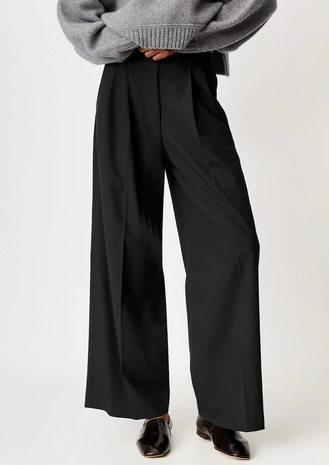 A person wearing black wide-leg pleated trousers with a gray knit sweater and dark brown shoes, shown from the waist down against a plain light background.