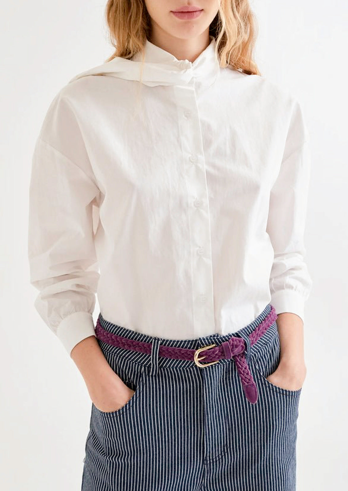 A woman wearing a white top, dark blue pinstriped jeans, and a deep purple braided belt.