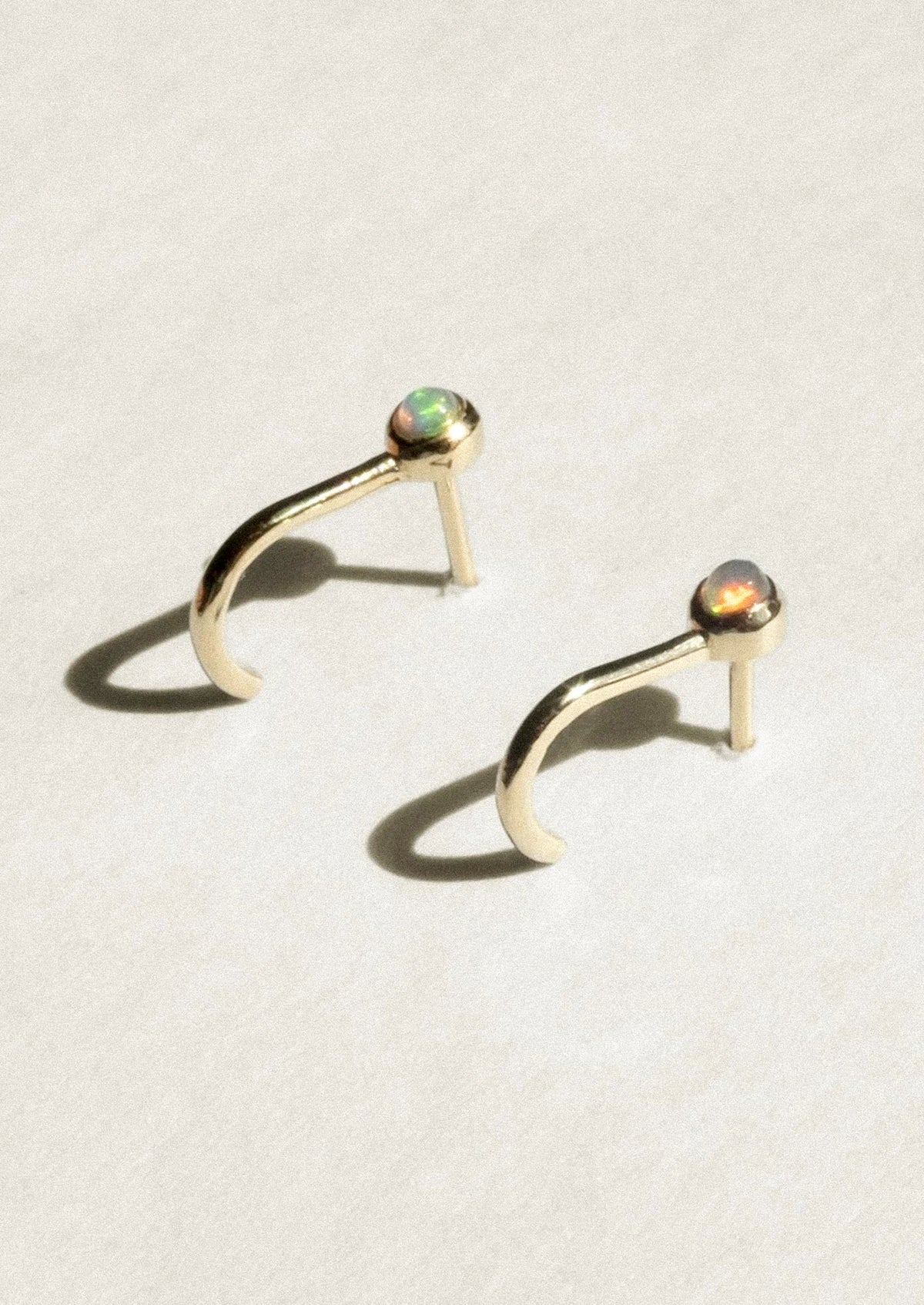 A pair of gold crescent-shaped hoop earrings with small round iridescent opal gemstones at the tips, displayed on a textured off-white surface with soft shadows.