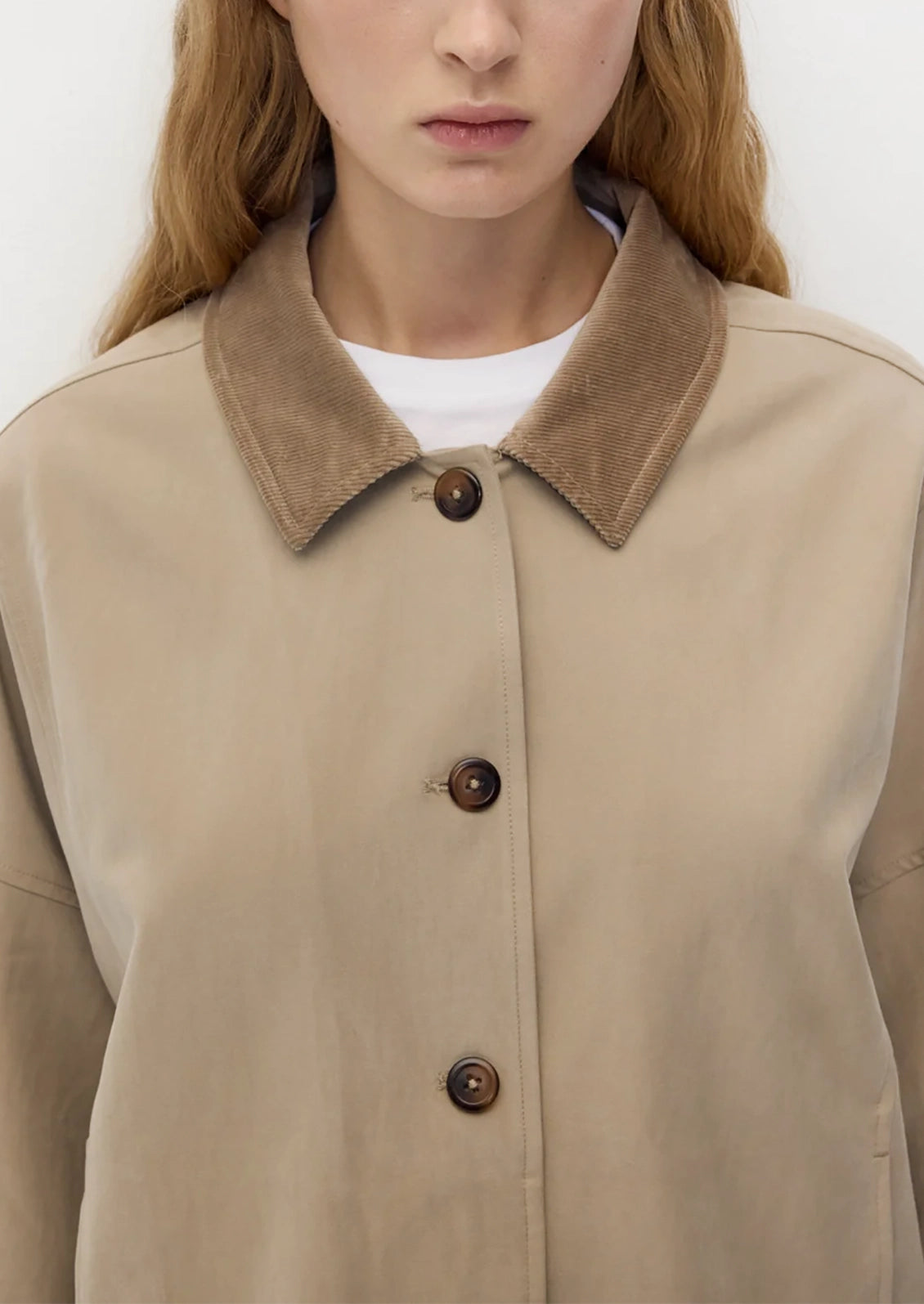 A close-up of a model wearing a beige utility-style jacket with a contrasting brown suede-like collar and dark brown buttons, layered over a white t-shirt.
