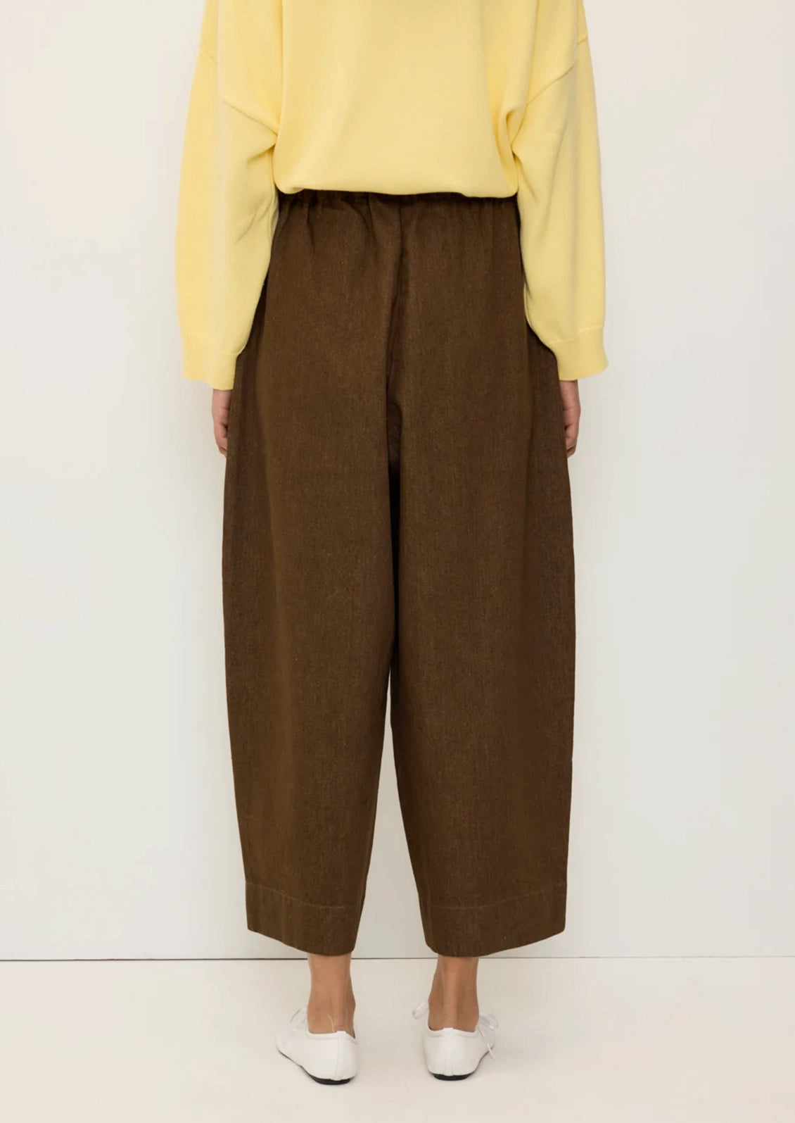 A person shown from the back wearing loose brown wide-leg cropped pants, a pale yellow long-sleeve top, and white shoes against a plain light background.