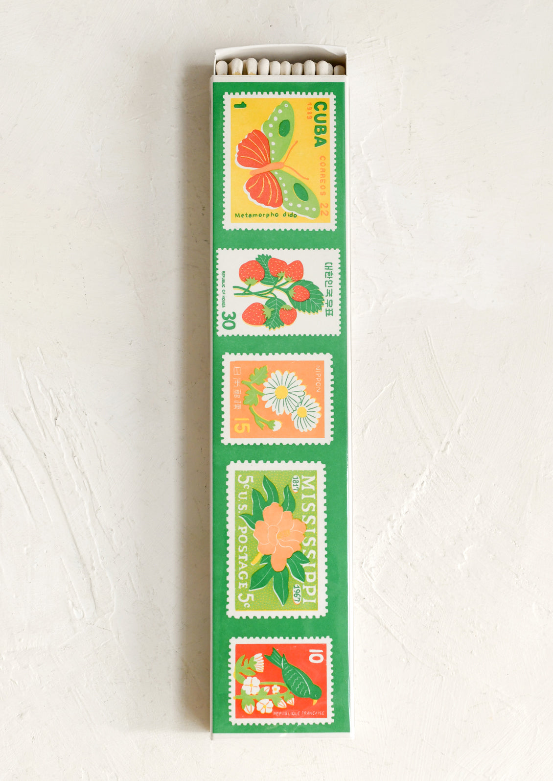 A long rectangular matchbox cover or decorative match holder with a green border and five vertical postage-stamp-style illustrations in yellow, red, orange, white, and green, shown standing upright on a white textured surface.