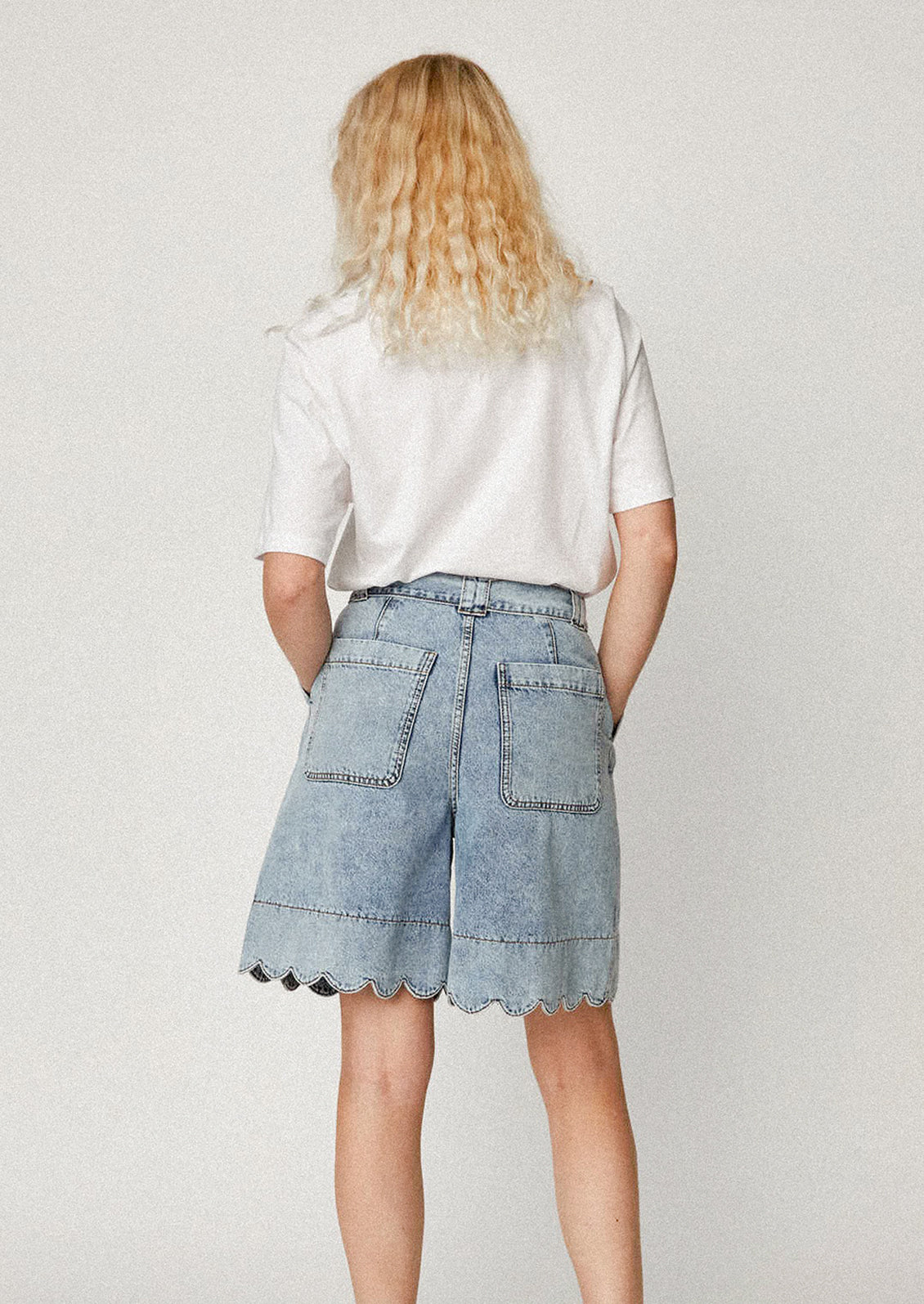 A person wearing a pair of medium blue wash bermuda shorts with scalloped hem.