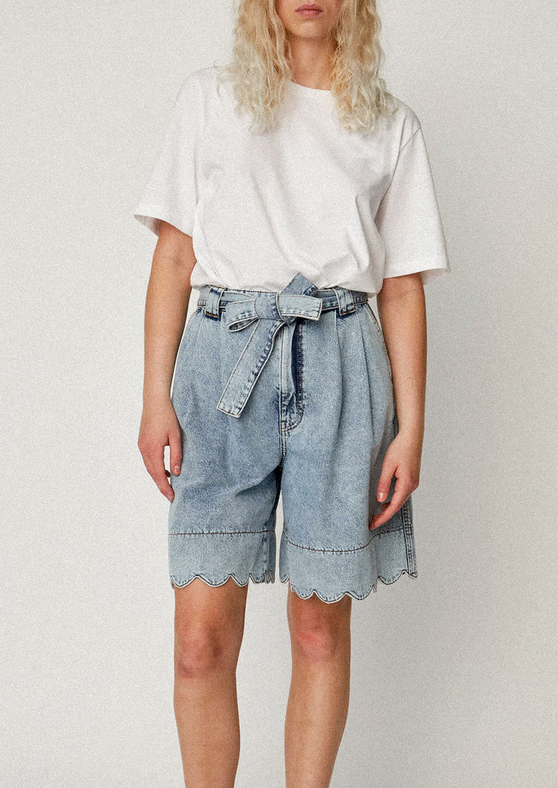 A person wearing a pair of medium blue wash bermuda shorts with scalloped hem.