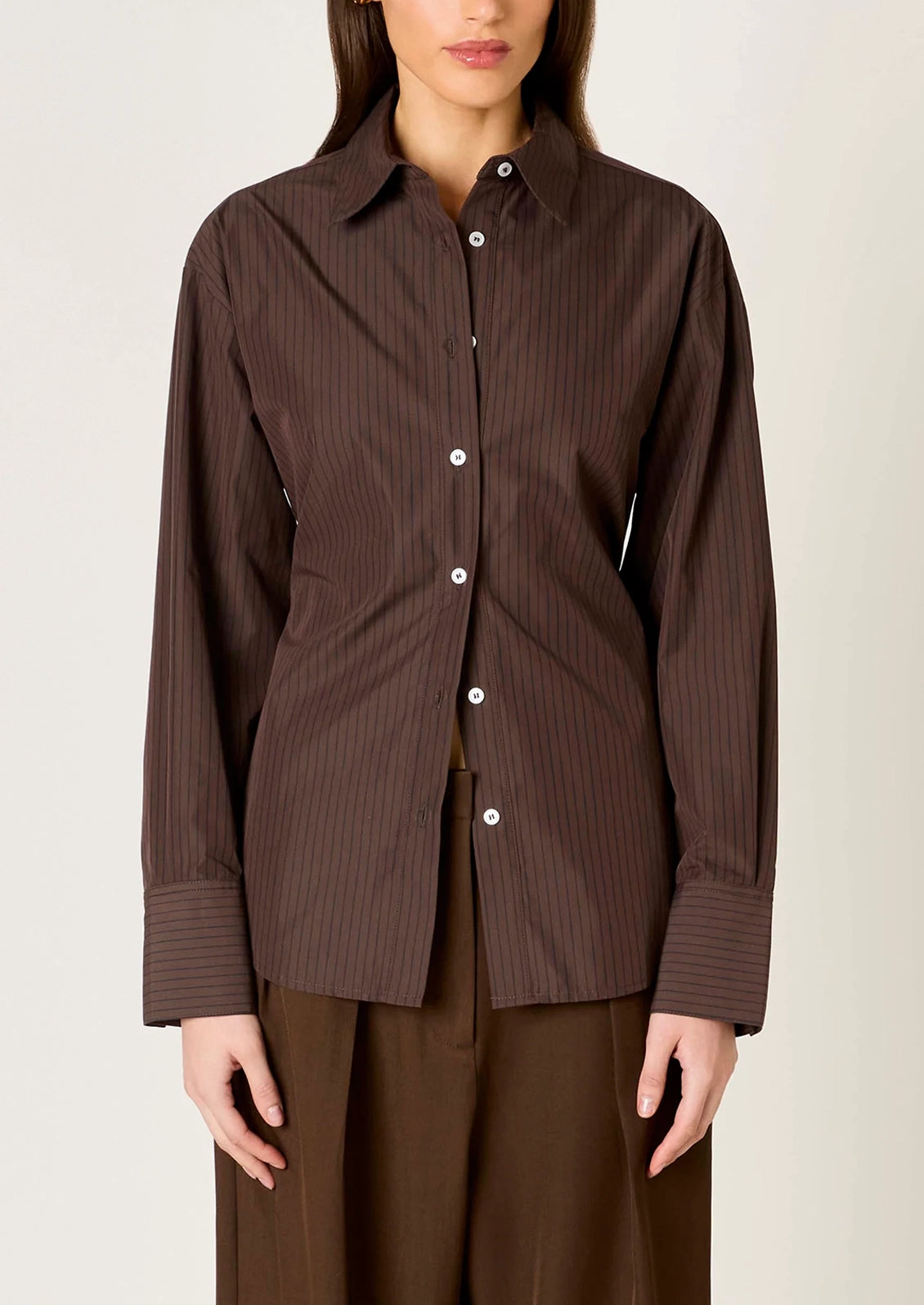 A woman wearing a dark brown long-sleeve button-up shirt with thin vertical pinstripes and white buttons, paired with matching brown trousers, against a plain light beige background.