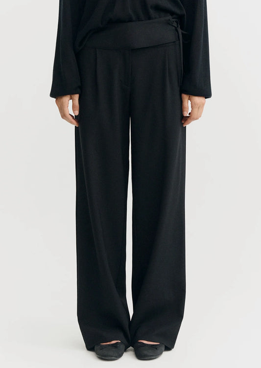 A model wearing black wide-leg trousers with a matching black top, shown from the waist down against a plain light gray studio background.