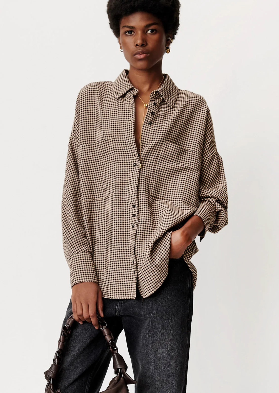 A woman modeling a brown and black checkered button-down shirt with dark gray jeans, holding a brown leather bag against a plain light gray background.