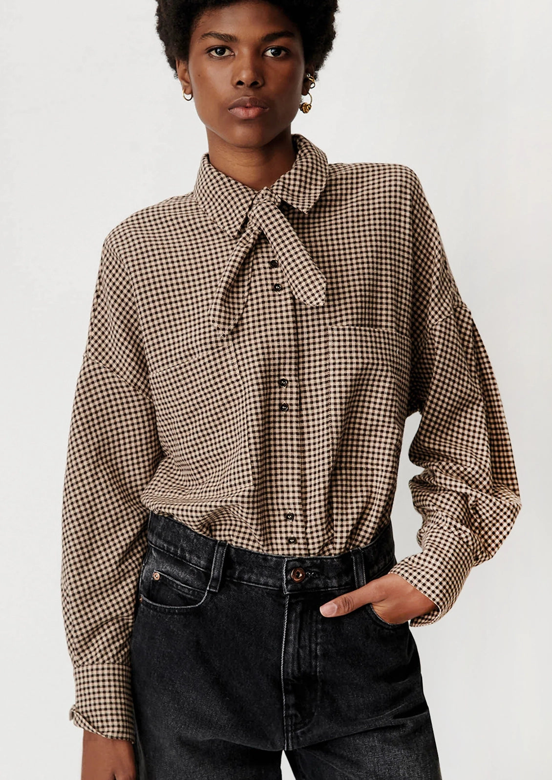 A woman wearing a brown and beige gingham long-sleeve button-up shirt with a tie-neck detail, paired with dark wash jeans, against a plain light background.
