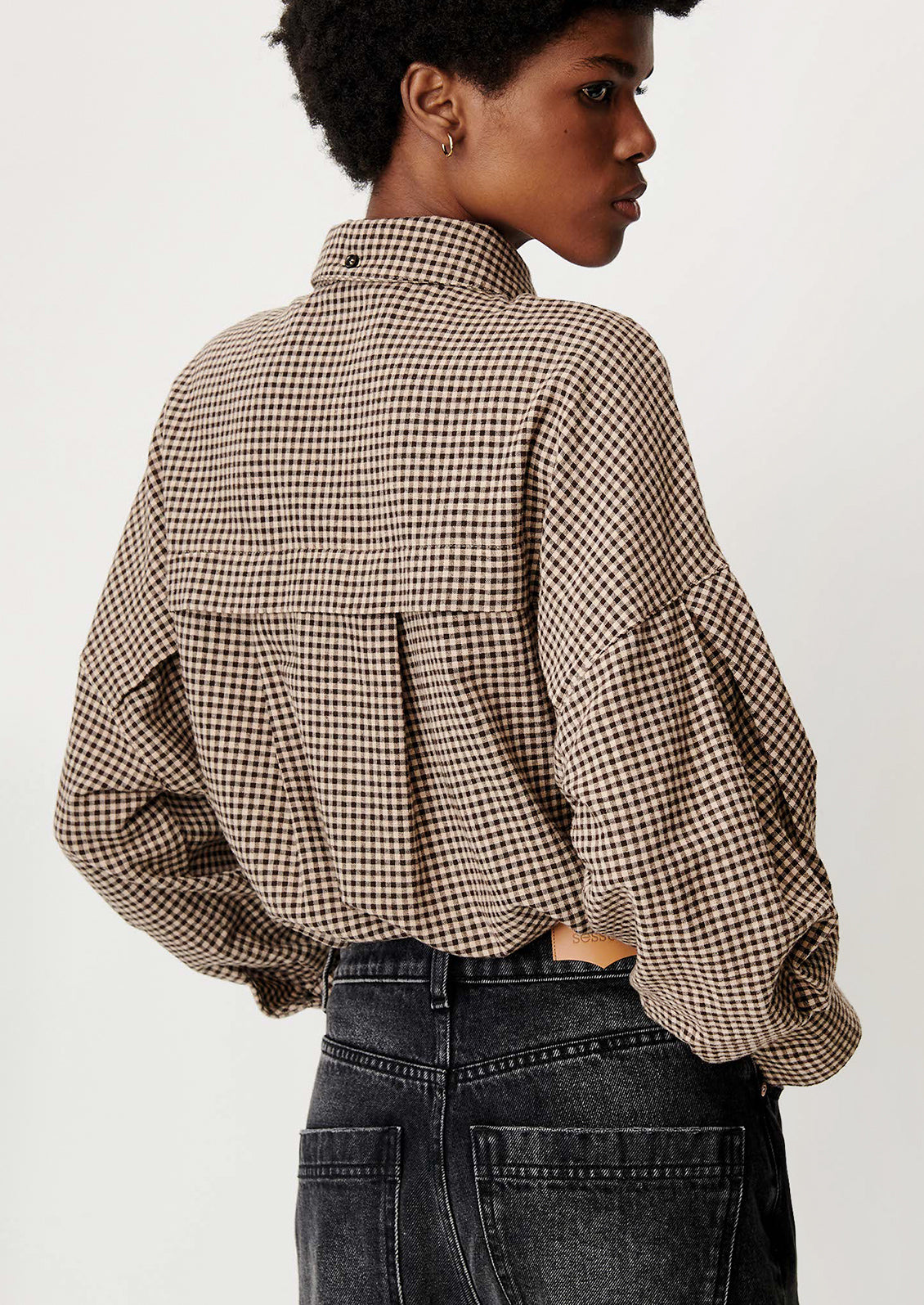 A model shown from the back wearing a brown and beige checkered long-sleeve blouse with a high collar and dark gray jeans, set against a plain light background.