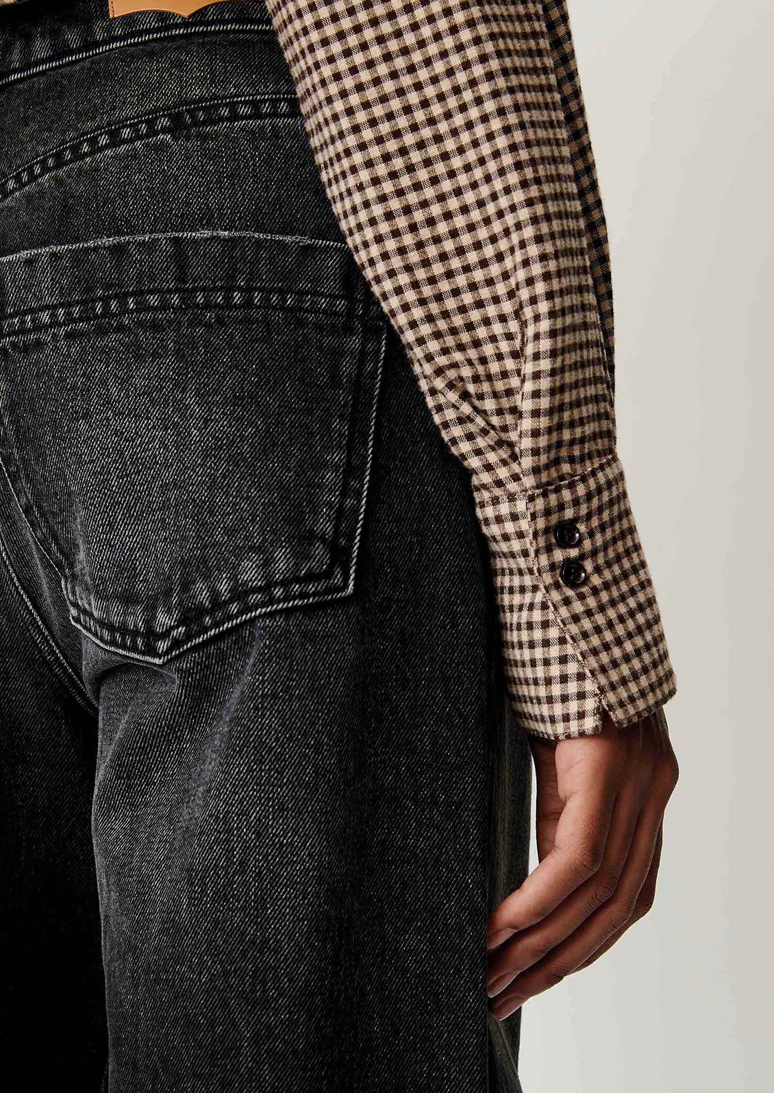 Close-up of a person wearing dark gray washed denim jeans and a brown and beige gingham long-sleeve shirt, focusing on the back pocket and cuff detail.