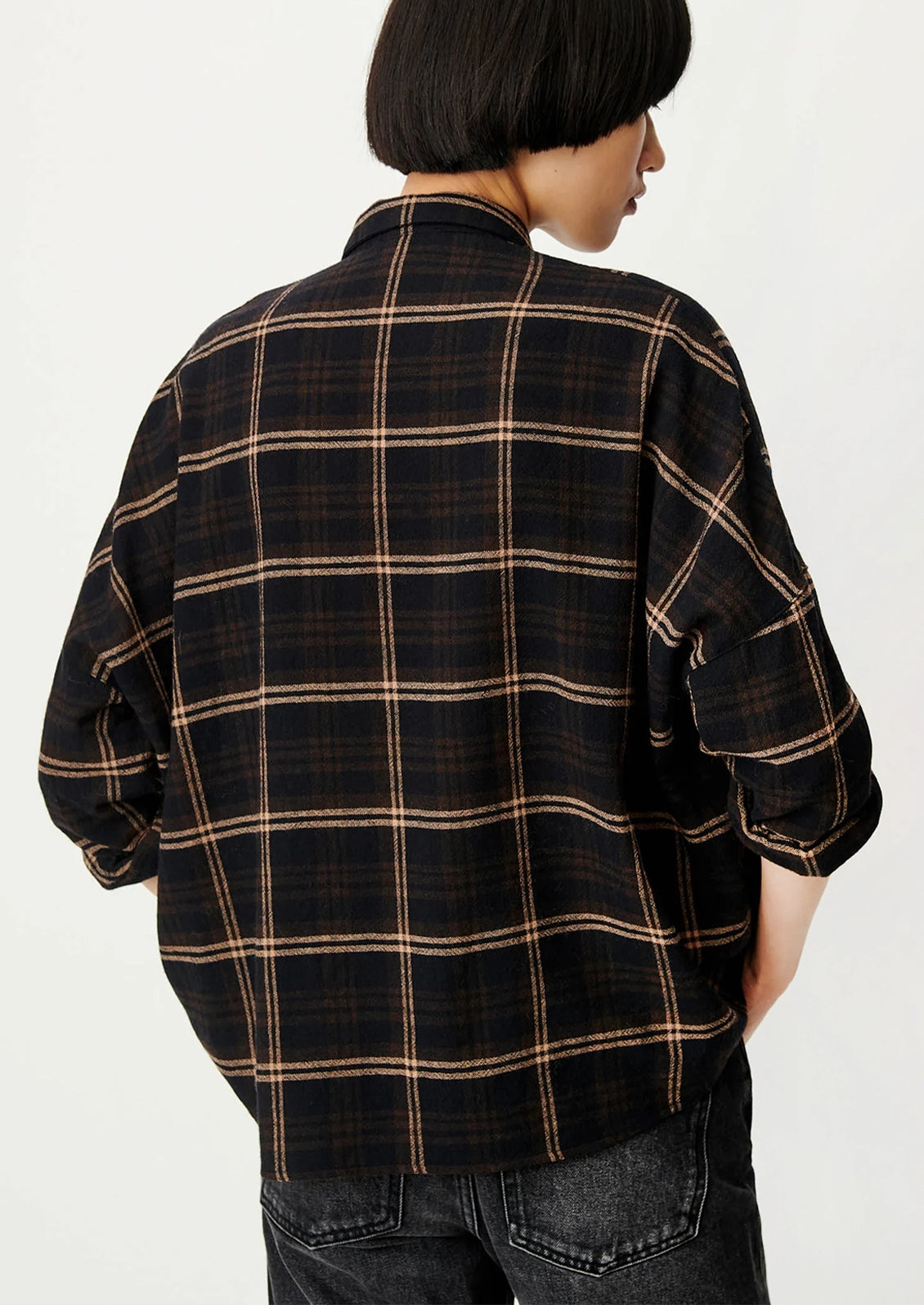 A woman wearing a black plaid button-down shirt with rolled-up sleeves and dark jeans, photographed against a plain light background.