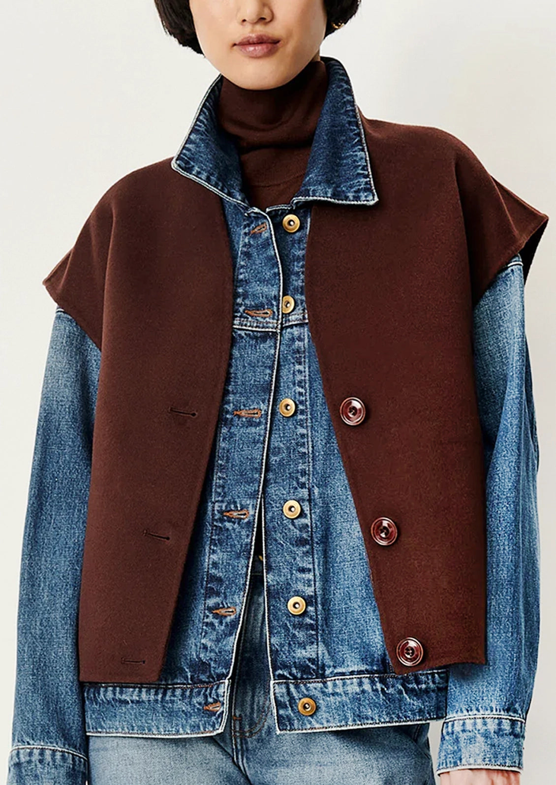 A model wearing a brown wool-like vest with denim sleeves over a blue denim jacket and a dark brown turtleneck, shot against a plain light background.
