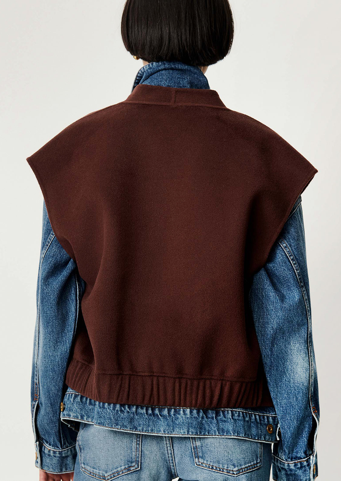 Rear view of a person wearing a sleeveless brown knit vest over a blue denim jacket and jeans against a plain light background.
