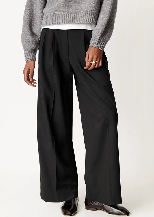 A person wearing black wide-leg pleated trousers, styled with a gray knit sweater, white undershirt, and dark brown leather shoes, shown from the chest down against a plain light background.