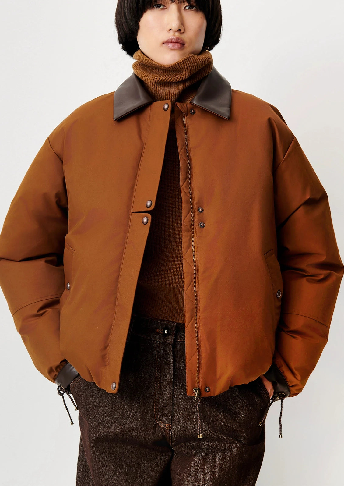 A model wearing a brown puffer-style jacket with dark brown leather-look collar panels, layered over a brown turtleneck sweater and dark brown pants, shot against a plain light gray studio background.