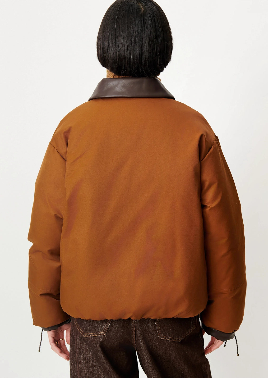 A person shown from the back wearing a cropped brown jacket with dark brown leather-like collar detail and dark brown pants, against a plain light background.