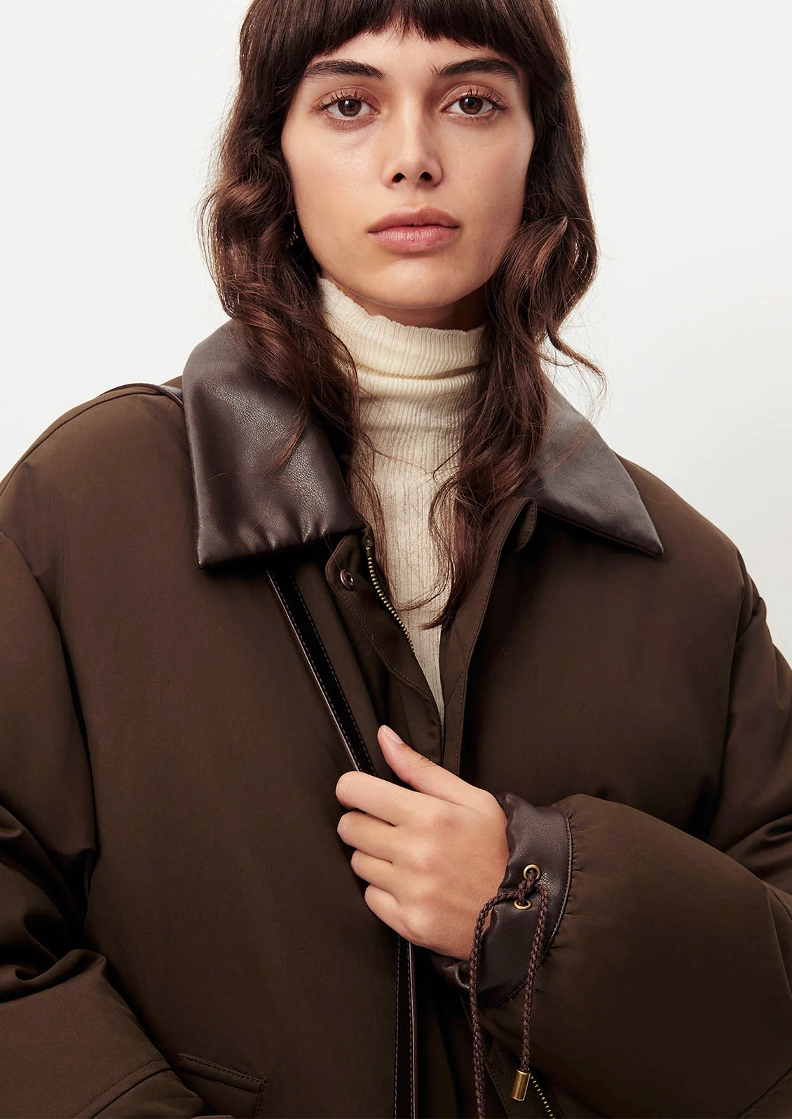 A woman wearing a dark brown puffer jacket with a wide dark brown collar over a cream turtleneck sweater, photographed against a plain light background.