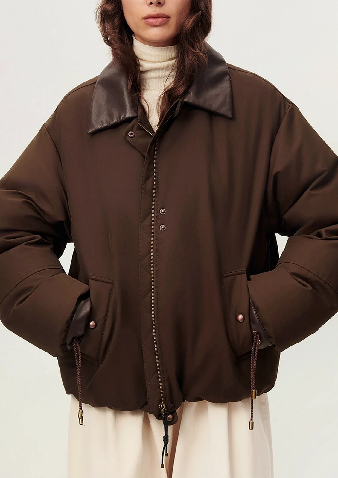 A model wearing an oversized dark brown puffer jacket with a contrasting dark brown collar, styled over a cream turtleneck and cream skirt against a light gray studio background.
