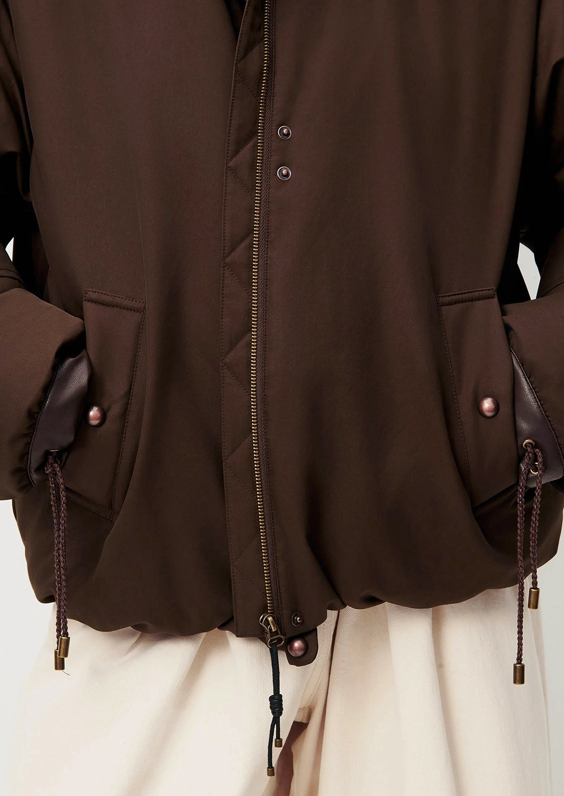 Close-up of a dark brown jacket with a front zipper, snap buttons, and drawstring details, worn over cream-colored pants.