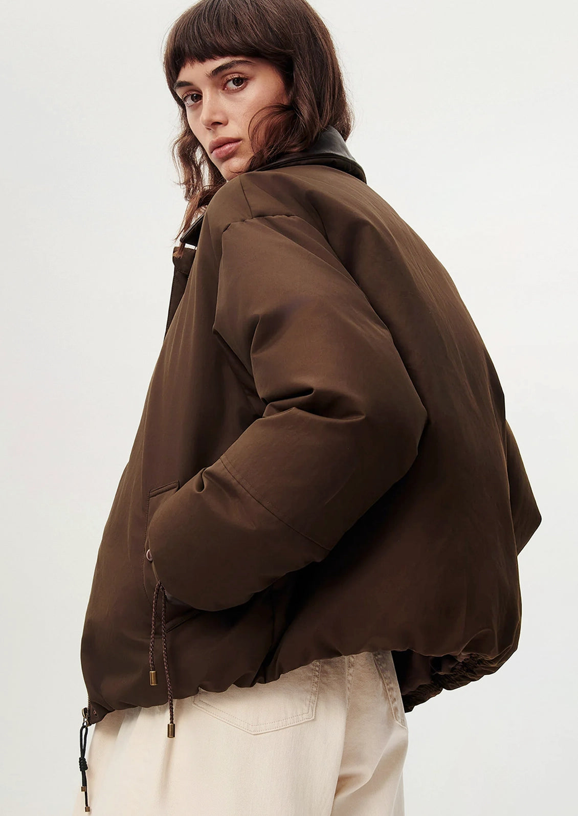 A model wearing a dark brown puffer jacket with a black collar, paired with cream-colored pants, photographed against a plain light gray background.