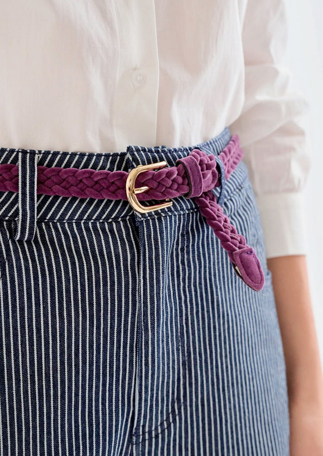 A person wearing a white top, dark blue pinstriped jeans, and a deep purple braided belt.