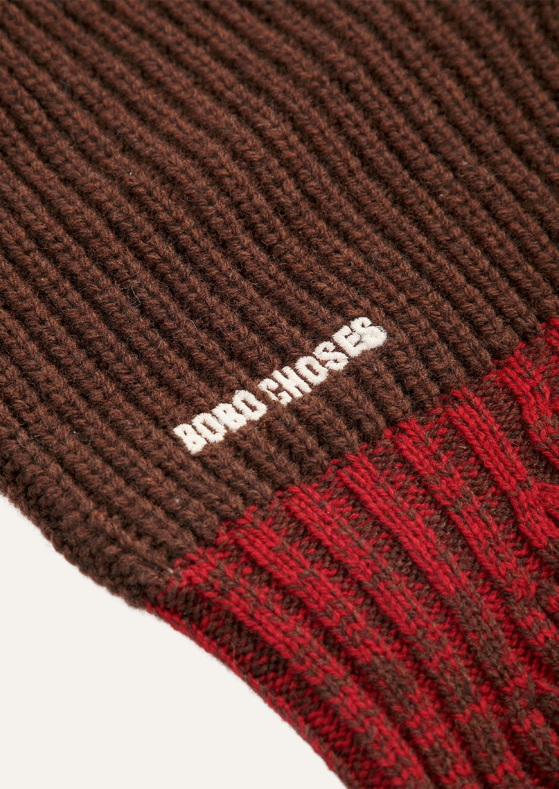 Closeup of a brown, red, and green color block balaclava with a ribbed knit design and the brand name "Bobo Choses" embroidered in white.