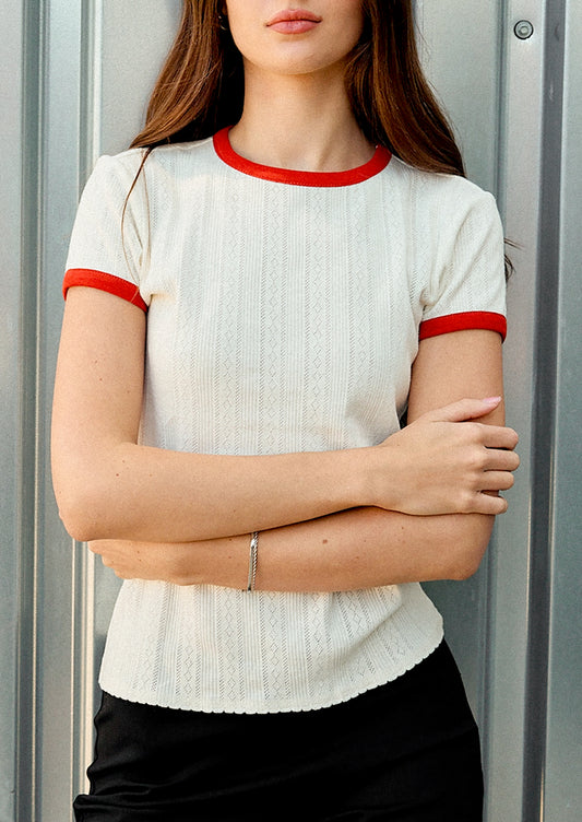 A woman wearing a short-sleeve ribbed knit top in off-white with red trim at the crew neck and sleeve cuffs, paired with black bottoms, standing against a gray corrugated metal wall.