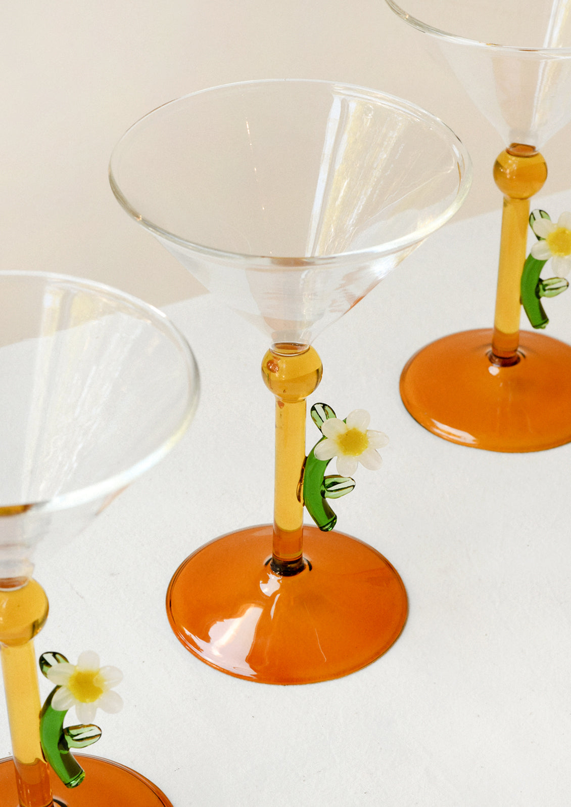 Clear glass martini glasses with orange-colored stems and bases, decorated with small white daisy and green leaf accents, arranged on a light surface.