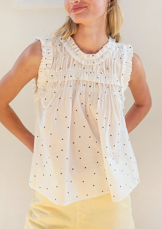 A woman wearing a sleeveless white blouse with black polka dots and a smocked ruffled neckline, paired with light yellow pants, against a plain beige background.