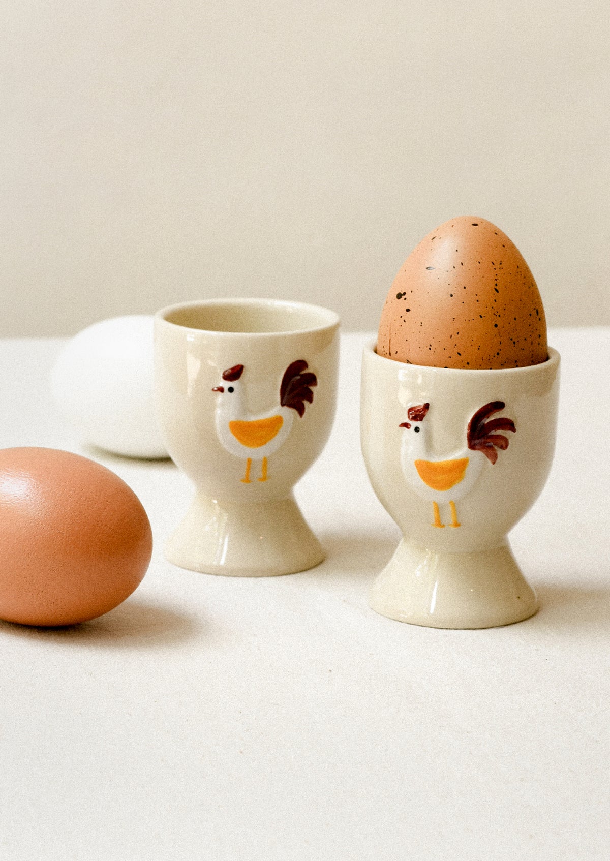 Two ceramic egg cups with chicken designs on a white surface, containing eggs.
