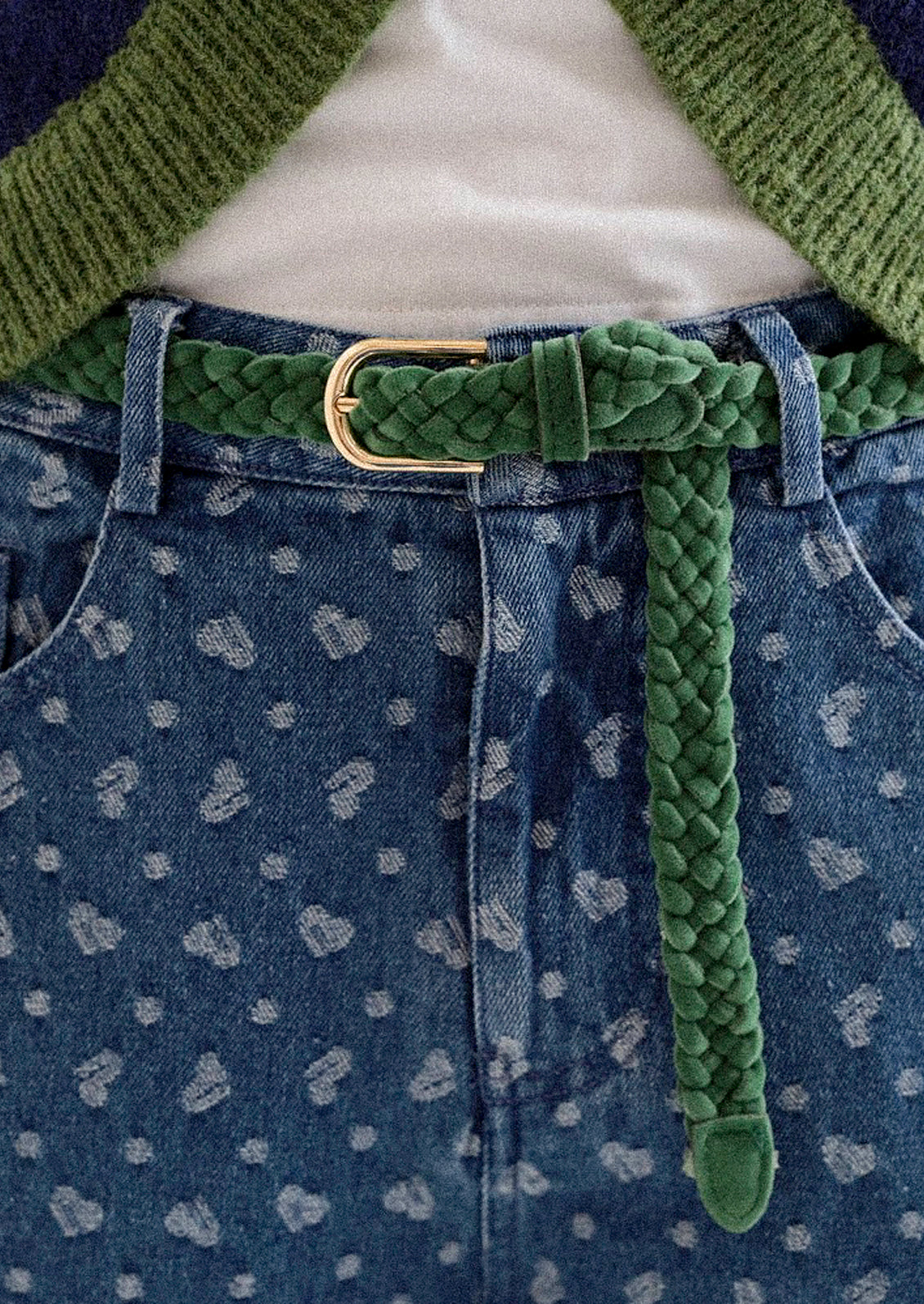 A person wearing a white top underneath a green and blue sweater, printed jeans, and a slim braided emerald green belt.