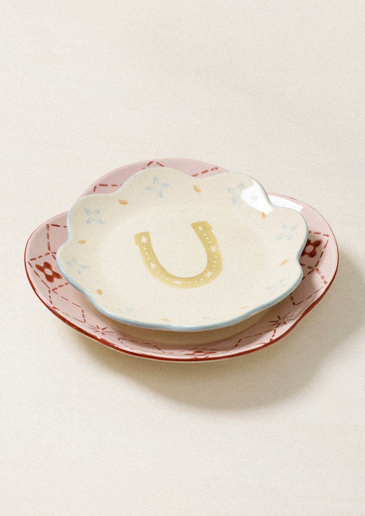 A small scalloped ceramic dish with a yellow horseshoe design in the center, nested on a larger pink and brown patterned plate against a light beige background.