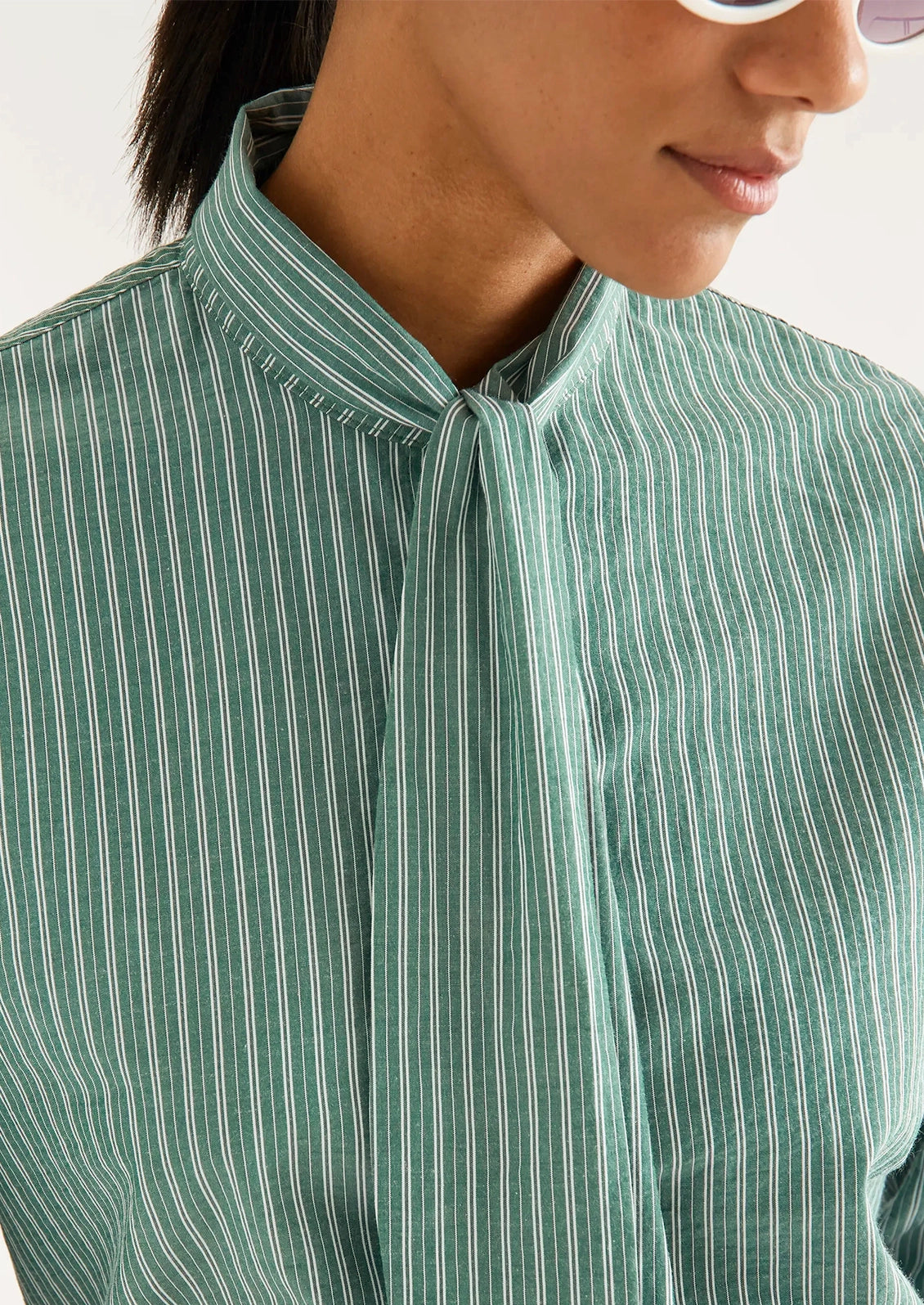 A woman wearing a long sleeve blouse with neck tie detail, in allover green and white stripe.