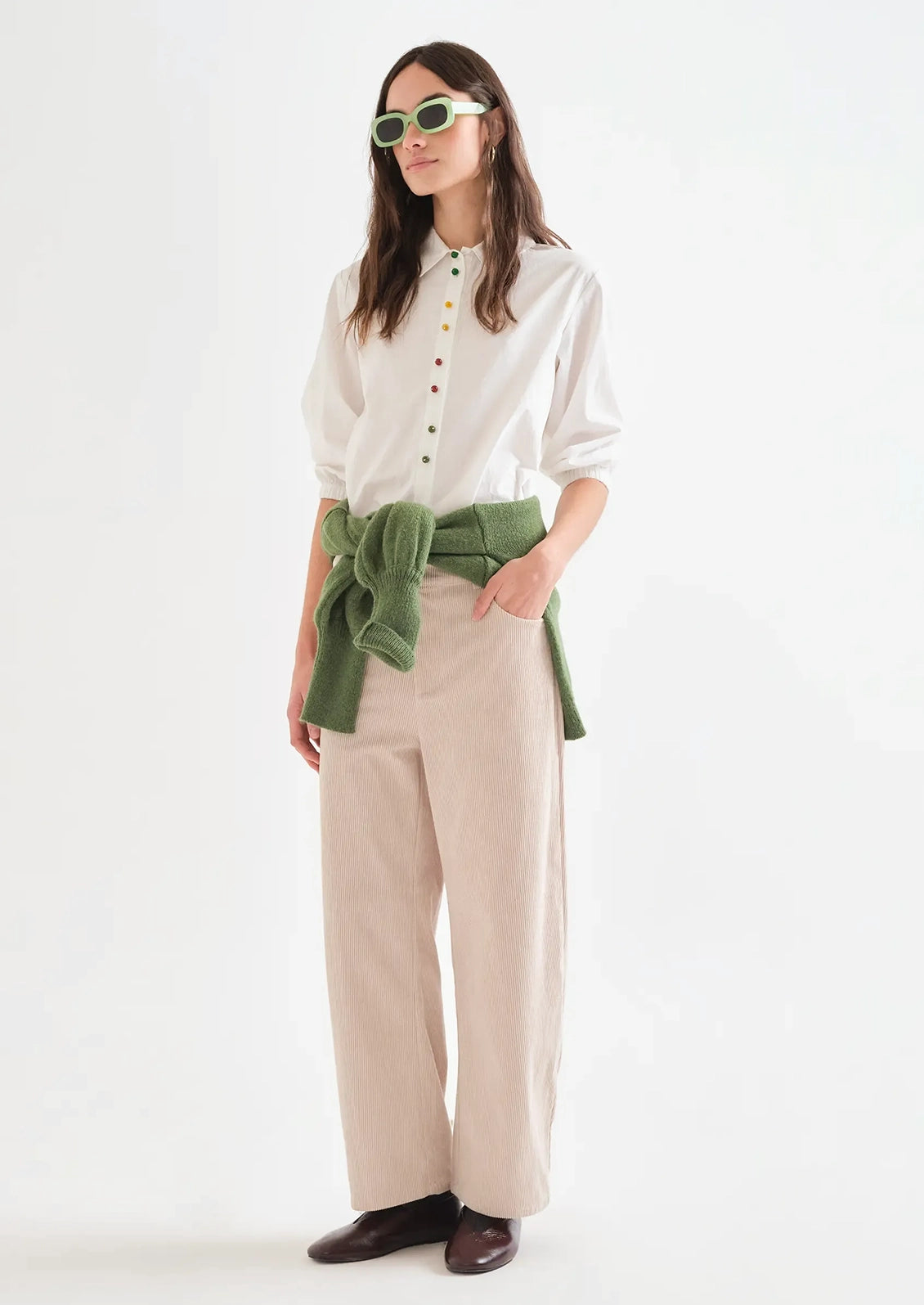 A woman modeling a white long-sleeve button-up shirt with colorful buttons, beige wide-leg trousers, an olive green knitted belt tied at the waist, and green-tinted sunglasses, standing against a plain white studio background.