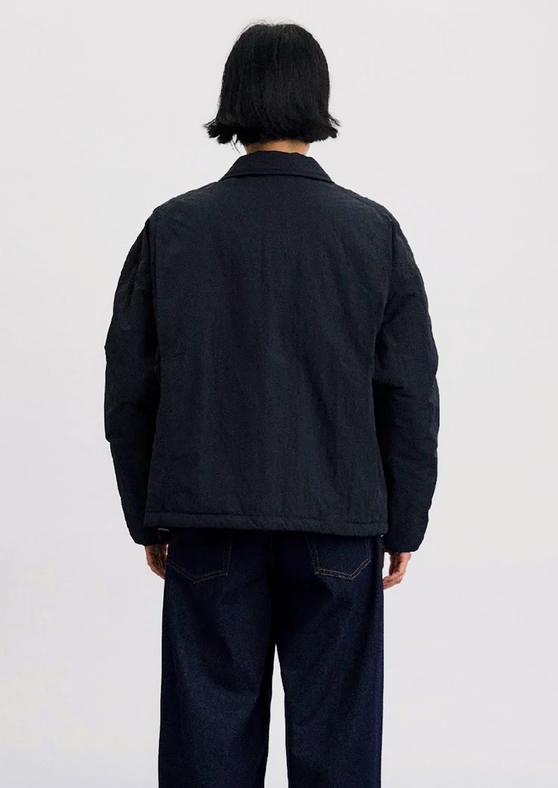 A person shown from the back wearing a dark navy quilted jacket and dark blue jeans against a plain light gray studio background.