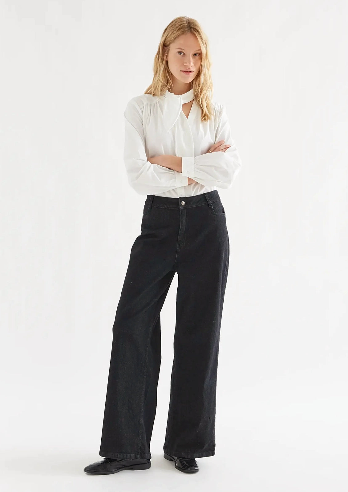 A woman modeling a white long-sleeve blouse with a high neck and black wide-leg jeans against a plain white studio background.
