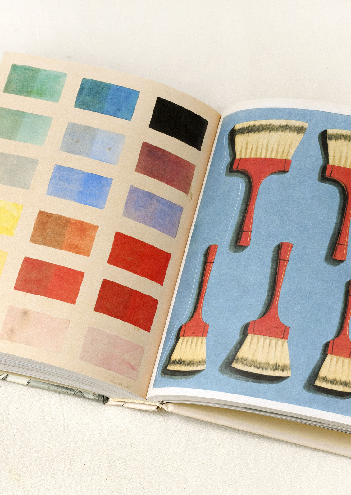 A sticker book sitting open on a table showing two pages, one with colorful, rectangular watercolor paint swatches and the other with various sizes paintbrushes with red handles on a blue background.