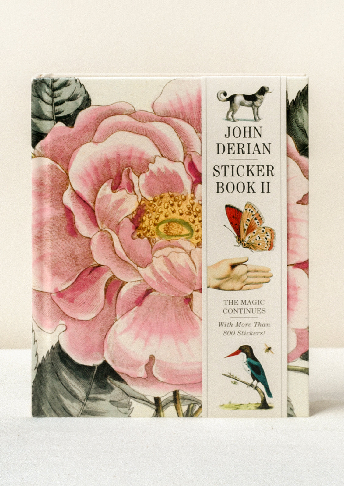 A sticker book standing on a table featuring a closeup of a pink flower on the cover and a paper band along the side with the title "John Derian Sticker Book II" and images of various animals.