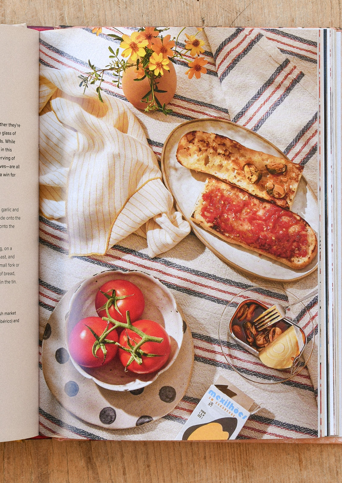 A book open on the table showing a picture of pan con tomate  a bowl of tomatoes, a vase with yellow flowers, and striped linens.