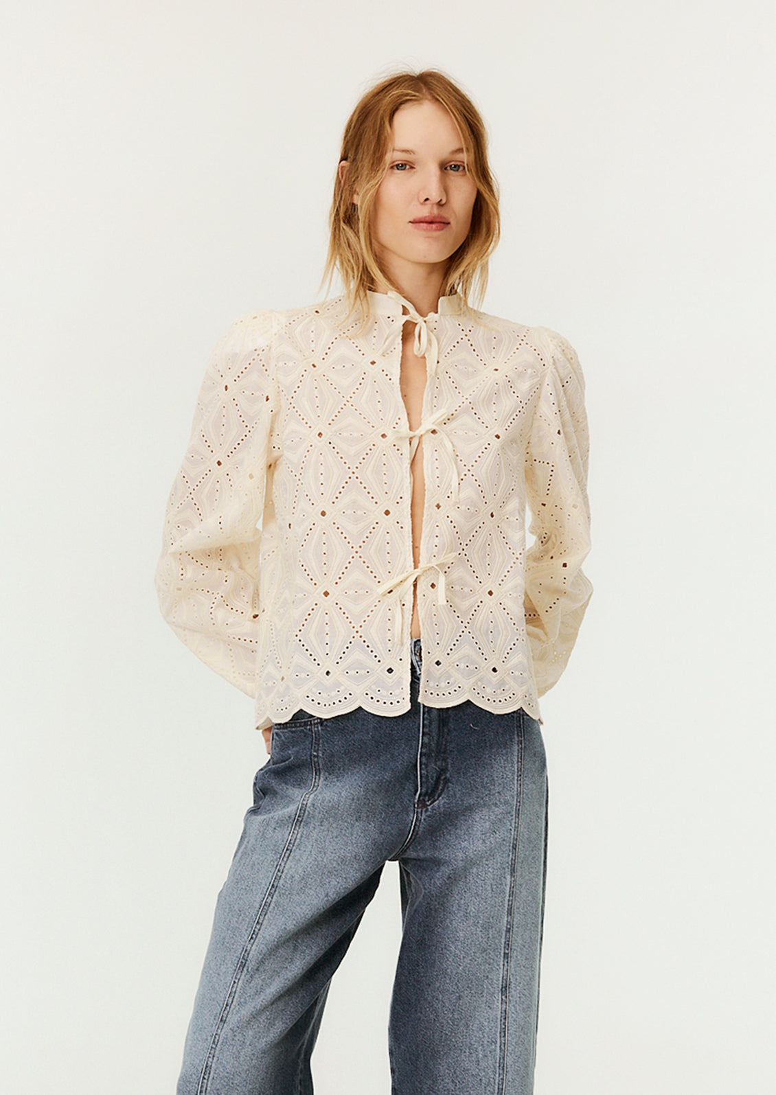 A woman wearing a cream lace blouse with mandarin collar and triple bow tie closure at front.