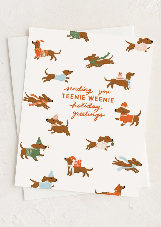 A white holiday greeting card featuring a playful illustration of dachshunds wearing colorful sweaters and scarves, with the text 'sending you TEENIE WEENIE holiday greetings' in orange and brown script. A second white card is partially visible underneath on a light textured background.