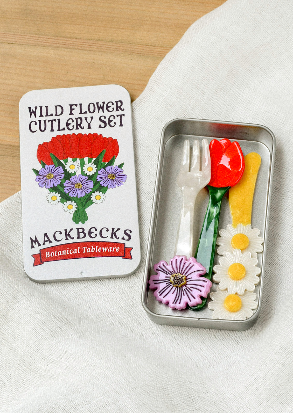 A small cutlery set in a silver tin that includes a fork, knife, and spoon. The fork has a purple flower at the end, the yellow knife has three daisies, and the spoon is in the shape of a red tulip with a green stem.