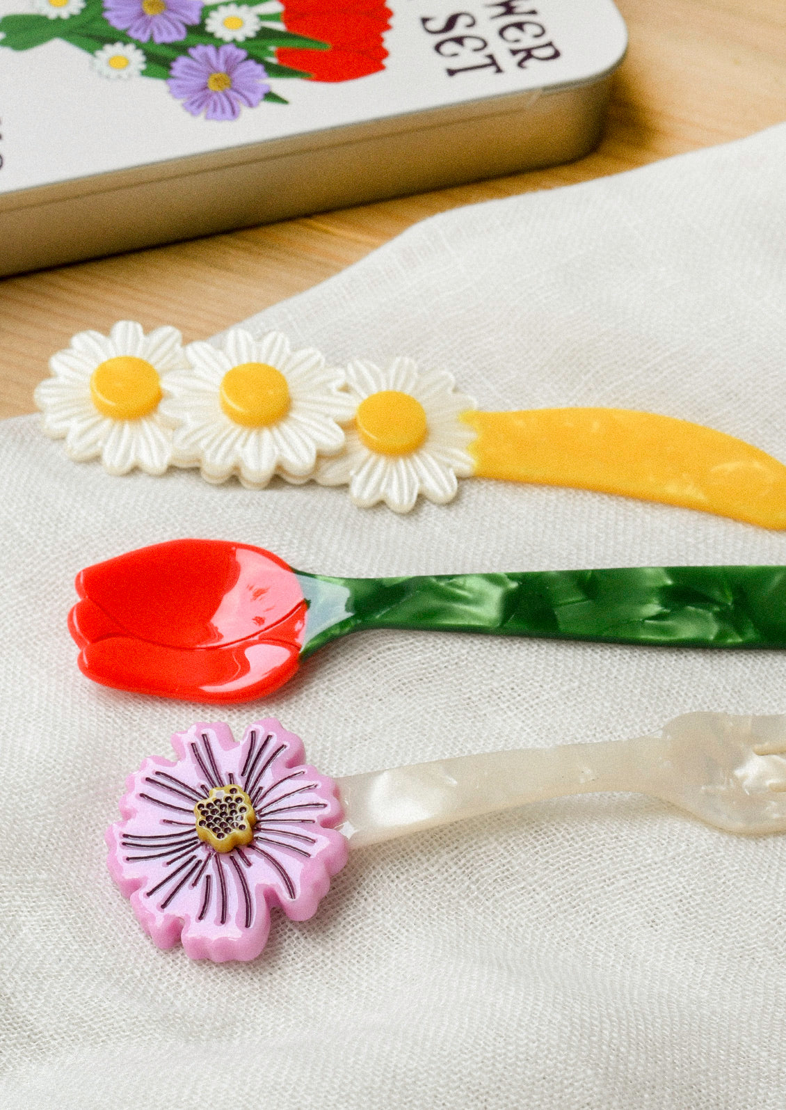 A small cutlery set in a silver tin that includes a fork, knife, and spoon. The fork has a purple flower at the end, the yellow knife has three daisies, and the spoon is in the shape of a red tulip with a green stem.