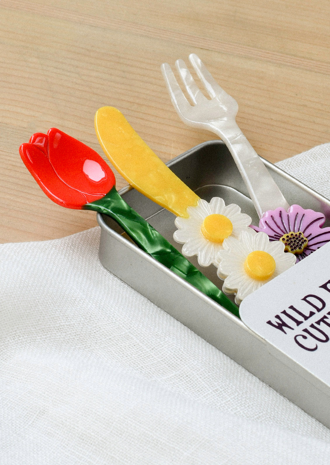 A small cutlery set in a silver tin that includes a fork, knife, and spoon. The fork has a purple flower at the end, the yellow knife has three daisies, and the spoon is in the shape of a red tulip with a green stem.