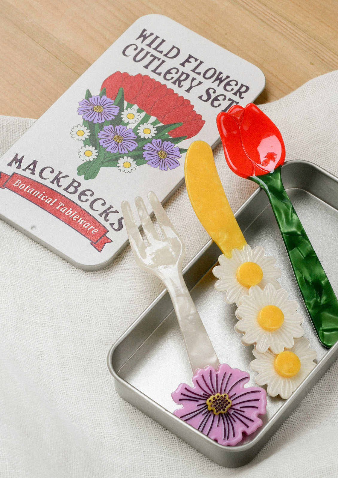 A small cutlery set in a silver tin that includes a fork, knife, and spoon. The fork has a purple flower at the end, the yellow knife has three daisies, and the spoon is in the shape of a red tulip with a green stem.