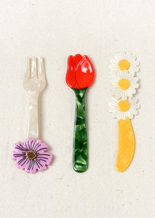 A small cutlery set that includes a fork, knife, and spoon. The fork has a purple flower at the end, the yellow knife has three daisies, and the spoon is in the shape of a red tulip with a green stem.