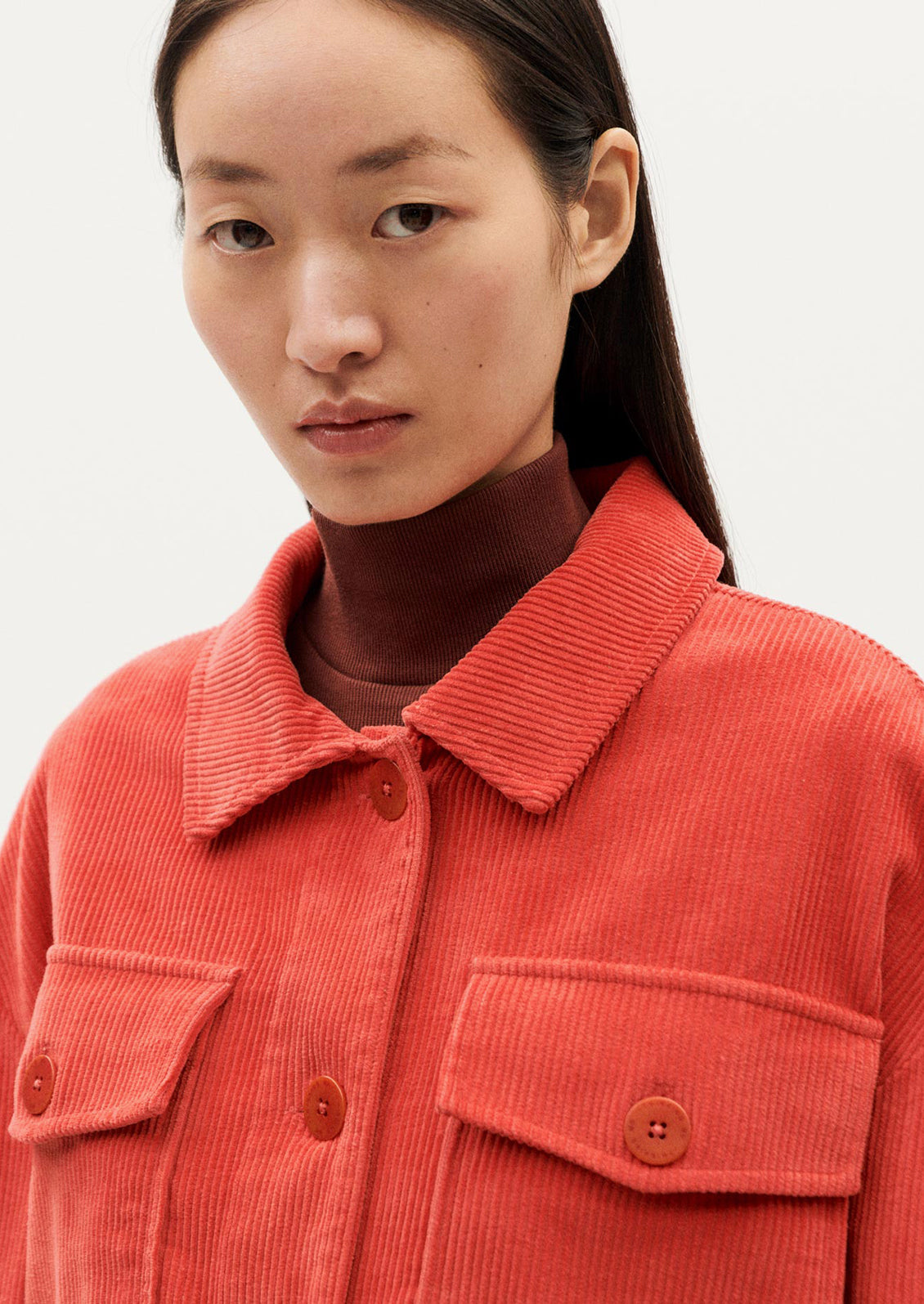 A woman wearing a shirt jacket style corduroy long sleeve jacket in red-orange hue.