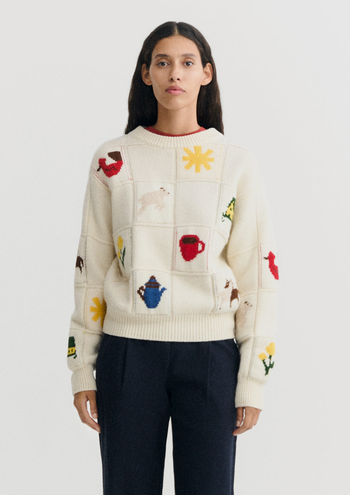 A model wearing a cream-colored knit sweater with colorful embroidered patches depicting animals, teapots, and flowers, paired with dark navy trousers, against a light gray background.