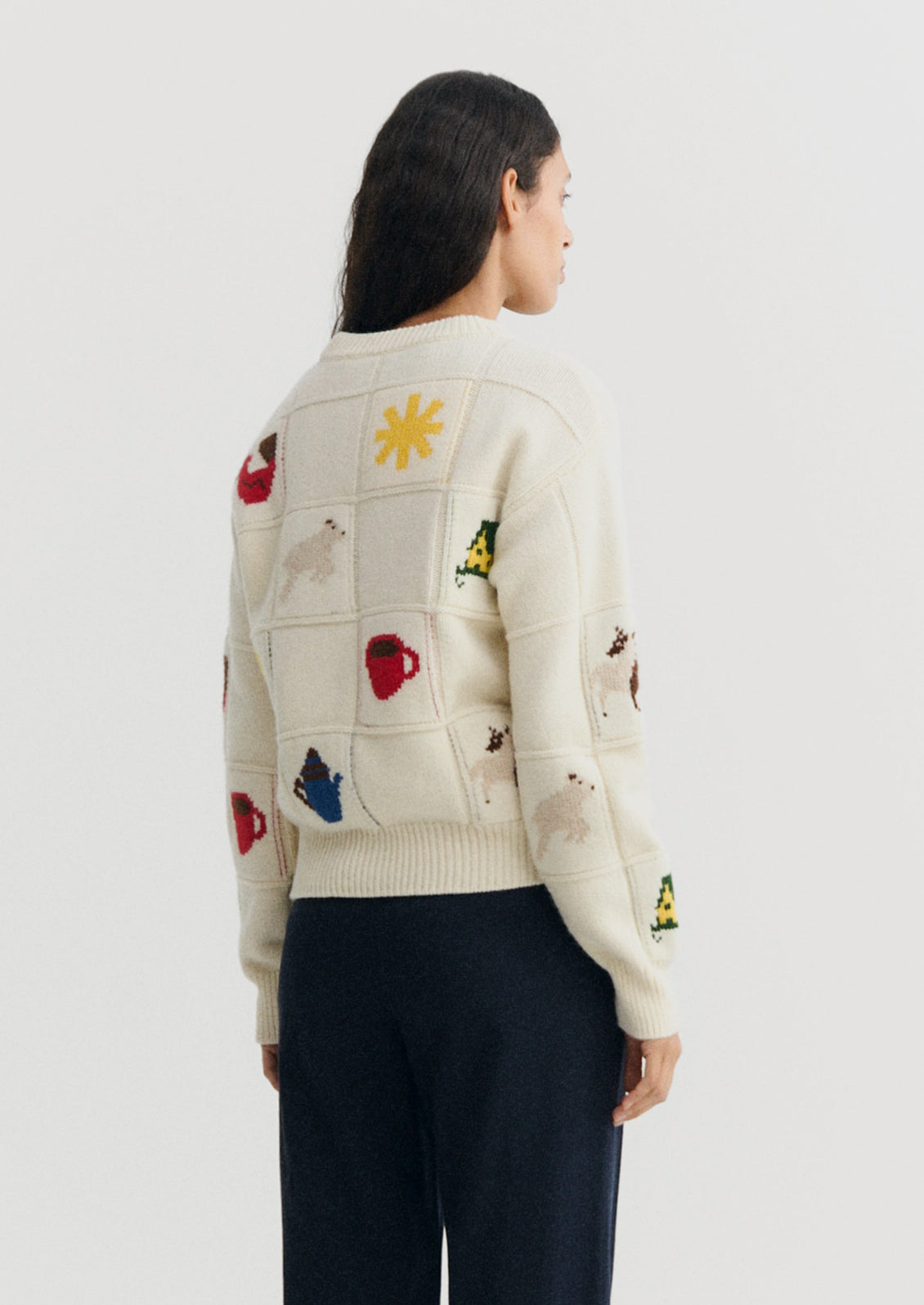 A model shown from the back wearing a cream-colored knit sweater with colorful knitted animal patches and dark navy pants, set against a plain light gray studio background.