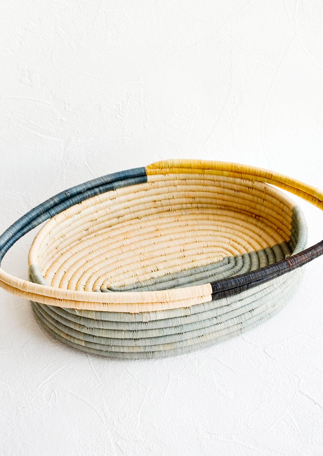 Oval bread basket in woven raffia with protruding handles at either side. Blue and yellow colorblock pattern.